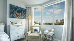 A bedroom with a bed, a chair, a nightstand, a dresser, a painting, a window with a city view, and a lamp.