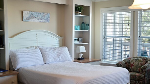 A bedroom with a bed, a nightstand, a lamp, a chair, a shelf with decorative items, and a window with blinds. - Cozy Gulf-View Studio Steps from the Beach (Bradenton Beach)