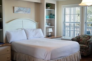 Un dormitorio con una cama, una mesita de noche, una lámpara, una silla, una estantería con adornos y una ventana con persianas.