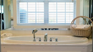 Un baño con una bañera grande, una ventana con persianas y una cesta de mimbre con toallas.