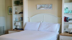 Una cama bien tendida con ropa de cama blanca, un cabecero blanco y una mesita de noche con una lámpara y libros.