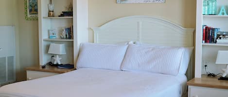 Ein ordentlich bezogenes Bett mit weißer Bettwäsche, einem weißen Kopfteil und einem Nachttisch mit Lampe und Büchern.
