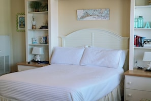 Una cama bien tendida con ropa de cama blanca, un cabecero blanco y una mesita de noche con una lámpara y libros.