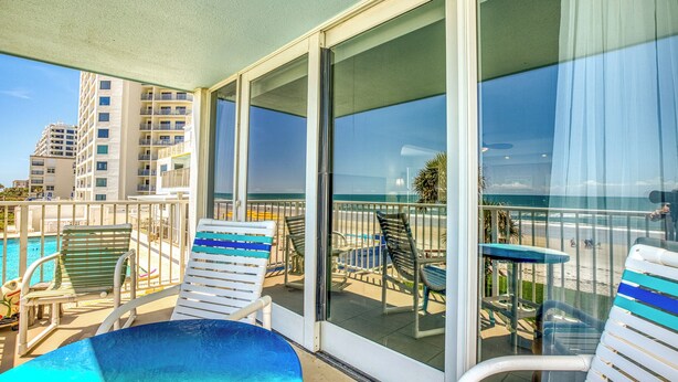 A balcony with a pool, lounge chairs, and a view of the beach and buildings.