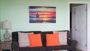 A living room with a sofa, two orange cushions, a painting of a sunset, a lamp, and a door. - Waterfront Views · One-Bedroom Bayfront Escape (Bradenton Beach)