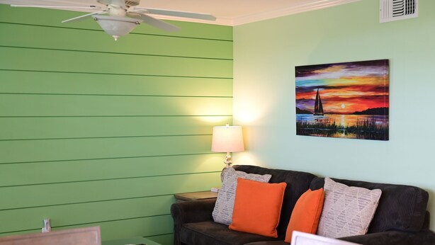 A room with a green wall, a black sofa with orange cushions, a small table with a lamp, and a painting of a sunset. - Waterfront Views · One-Bedroom Bayfront Escape (Bradenton Beach)