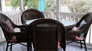 Une table et des chaises en osier sur une véranda dotée d’une balustrade, une table en verre et une vue sur les arbres et les bâtiments.