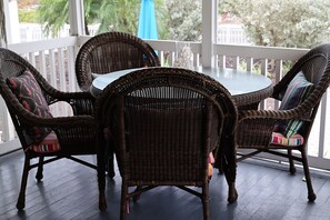 Een tafeltje en stoelen van riet op een veranda met een balustrade, een glazen tafel en uitzicht op bomen en gebouwen.
