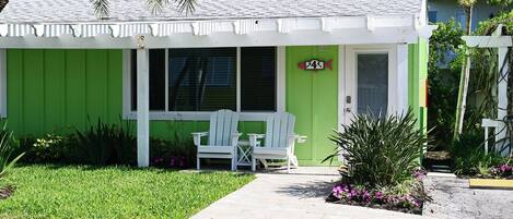 A small, single-story building with a green exterior and white trim. There is a small porch with two white chairs and a table.