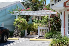 A parking area with a white pergola, surrounded by lush greenery and pink flowers.