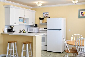 A compact kitchen with white cabinetry, a refrigerator, a microwave, and a coffee maker. There are two stools and a dining table with chairs. - Quiet Island Comfort · Cozy 1 bedroom Retreat (Bradenton Beach)