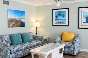 A room with a light blue wall, a ceiling fan, a sofa with floral upholstery, a small table with a map, and framed beach-themed artwork.