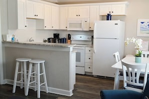 Una cocina con armarios blancos, un refrigerador blanco, un microondas, una estufa y un lavabo. Hay dos taburetes blancos y una mesa con sillas.