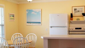 Une cuisine avec un réfrigérateur blanc, un coin repas avec des chaises blanches et un tableau sur le thème de la plage accroché au mur.