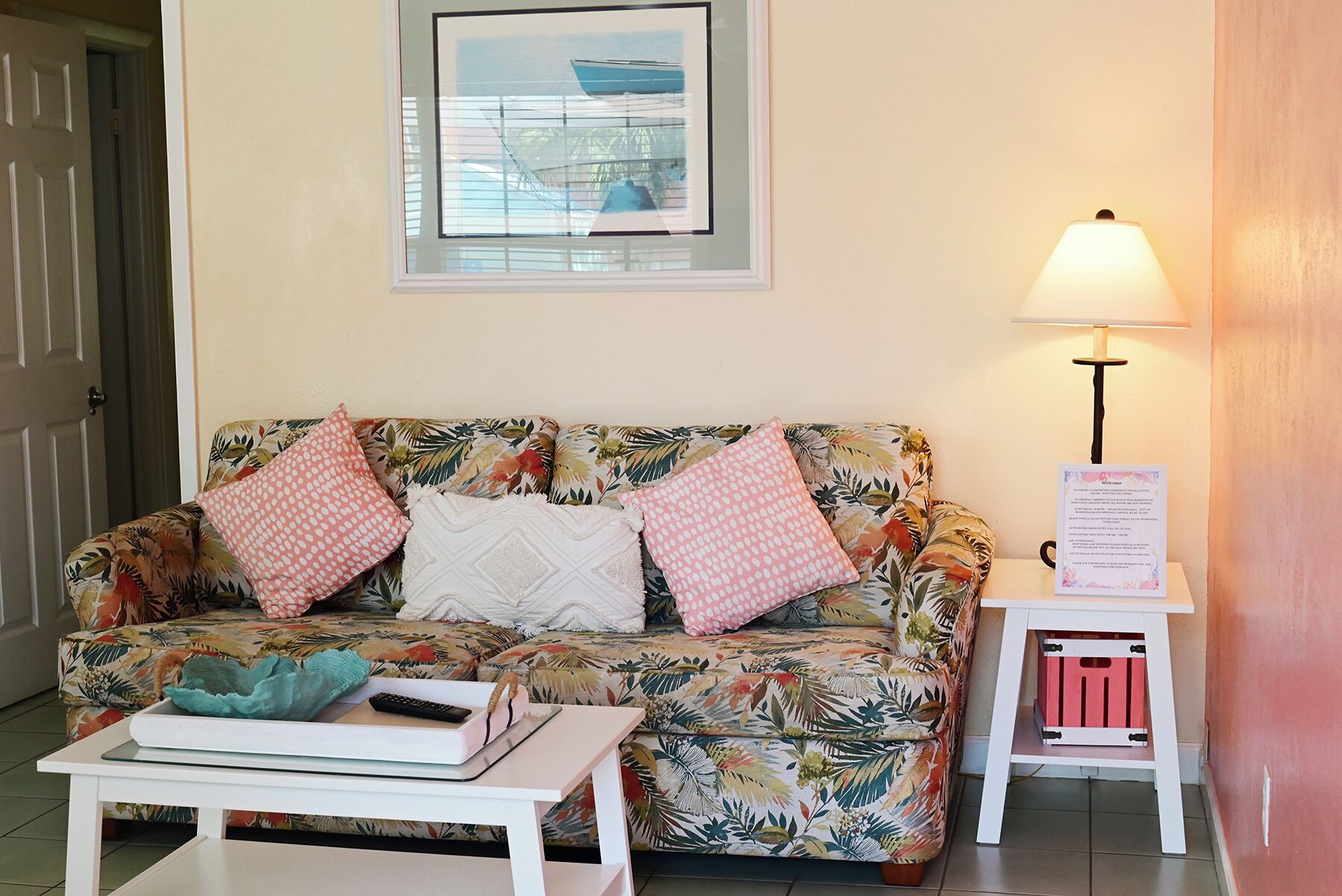 A room with a floral-patterned sofa, a small white table with a remote control, a lamp, and a framed picture on the wall.