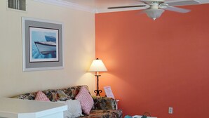 Une pièce avec un ventilateur de plafond blanc, un tableau encadré représentant un bateau, un canapé à motifs floraux, une table avec une lampe et un mur rouge.