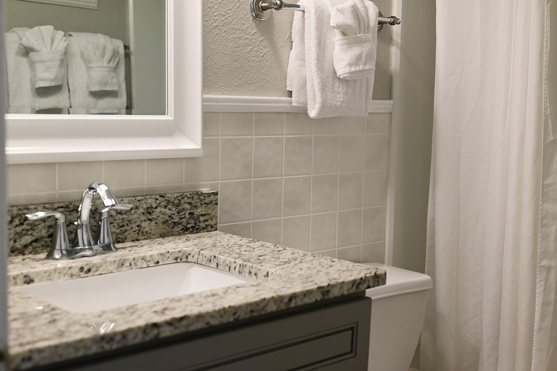 Un baño con lavamanos de encimera de granito, un espejo y un perchero con toallas blancas.