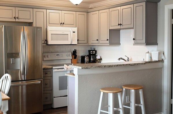 A kitchen with white cabinets, stainless steel appliances, a microwave, a coffee maker, and two bar stools.
