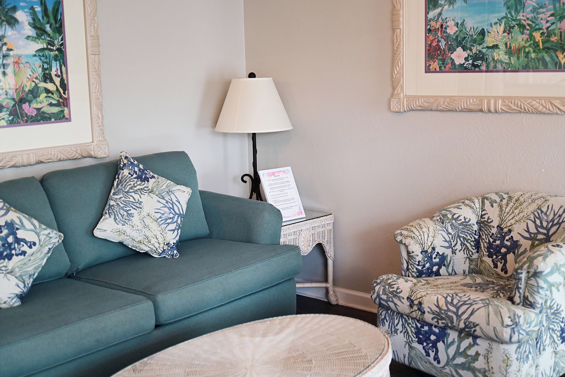 Una sala de estar con un sofá color turquesa, un sillón estampado floral, una lámpara y dos cuadros con motivos tropicales.