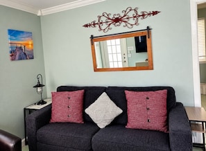 A dark-colored sofa with three pillows, a small table with a lamp, a wall-mounted mirror, and a framed picture on the wall. - Upgraded Coastal Style · Spacious South-Side 1 bedroom (Bradenton Beach)