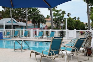A pool area with lounge chairs and umbrellas, surrounded by a white fence and palm trees. - Upgraded Coastal Style · Spacious South-Side 1 bedroom (Bradenton Beach)