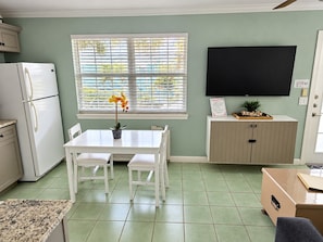 A kitchen with a dining table, chairs, a refrigerator, a TV mounted on the wall, and a window with blinds. - Upgraded Coastal Style · Spacious South-Side 1 bedroom (Bradenton Beach)