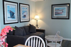 A room with a grey sofa, a small round table with a lamp, and three framed pictures of boats on the wall.
