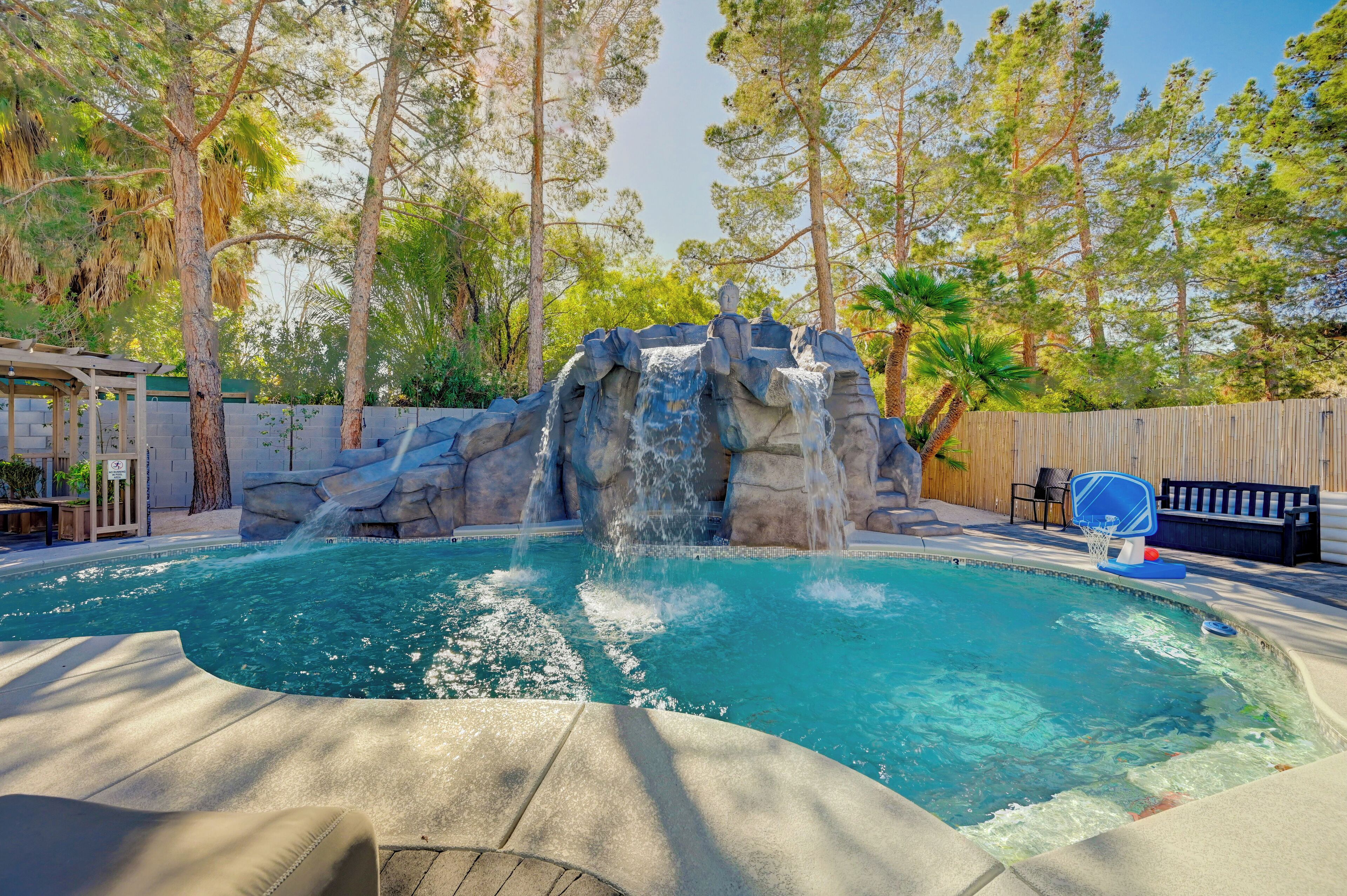 Piscina com cascata, cercada por árvores e um muro de madeira.