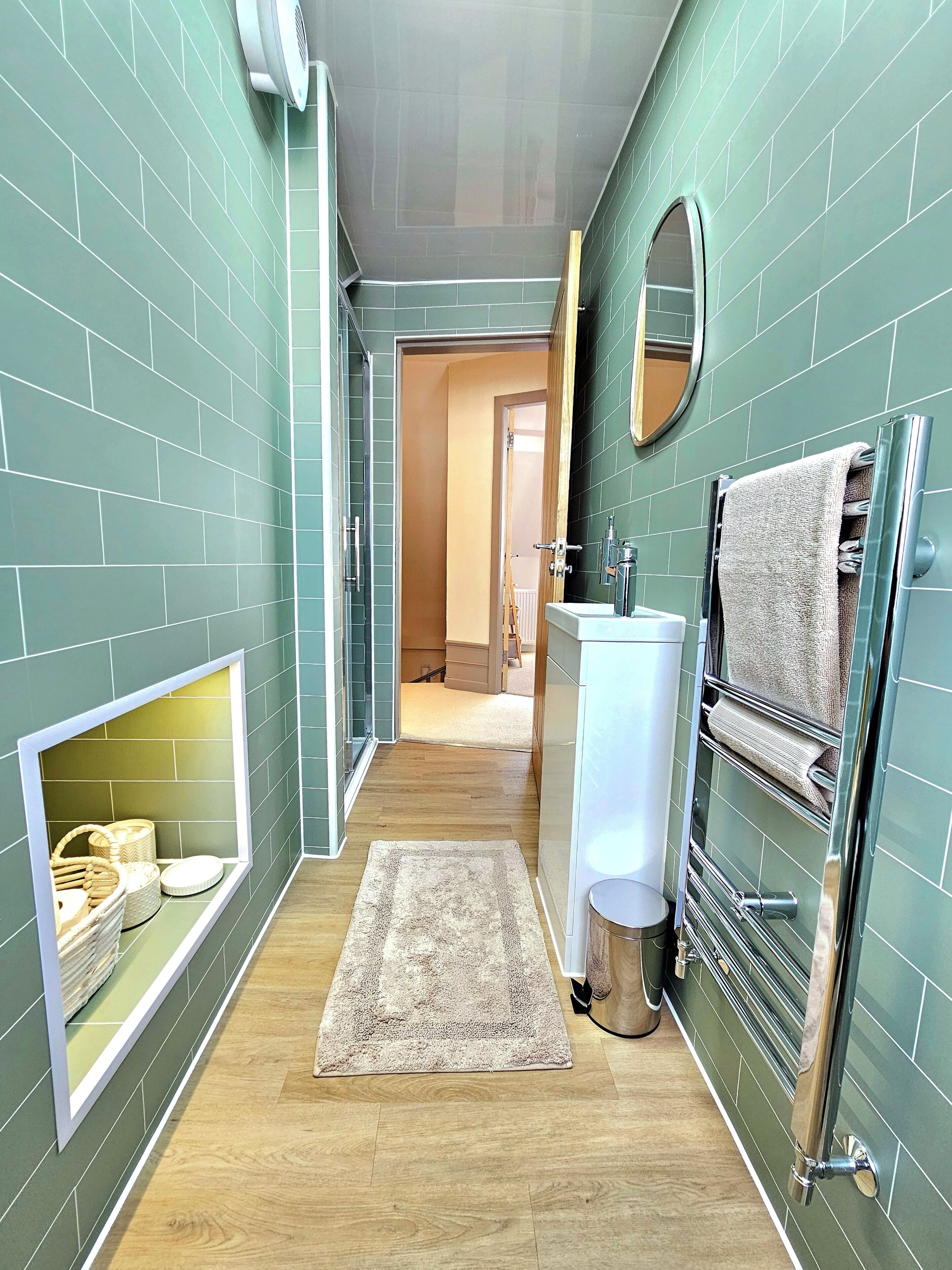 Un estrecho pasillo con paredes de azulejos verdes, un lavamanos blanco, un perchero y una puerta de madera que da a otra habitación.