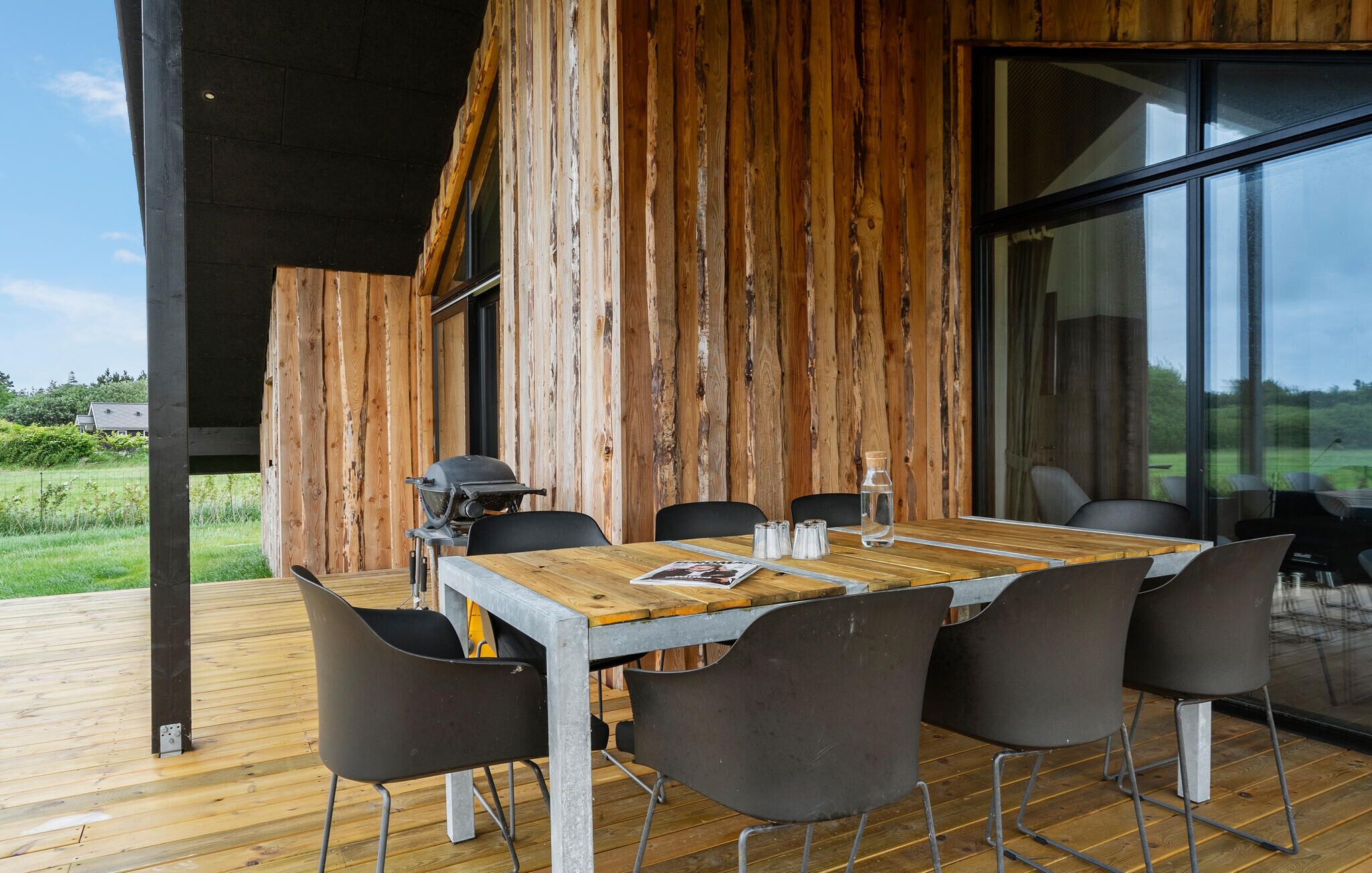 An outdoor dining area with a wooden table and chairs, a grill, and a glass wall offering a view of the outdoors.