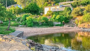 A riverside view with a wooden dock, rocky shoreline, and a house with a balcony overlooking the water. - Nice home in Klokkarstua with WiFi (Klokkarstua)