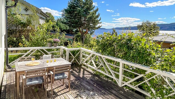 A wooden deck with a table and chairs, surrounded by lush greenery and a view of a lake. - Nice home in Klokkarstua with WiFi (Klokkarstua)