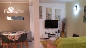 A modern living room with a sofa, a dining table with chairs, a flat-screen TV, and decorative wall art. - Large T2 ground floor In the countryside of Fort de France (Fort-de-France)