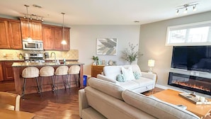Una sala de estar moderna con un sofá, una mesa de centro y un televisor, conectada a una cocina con armarios de madera y una zona de comedor con sillas.