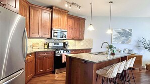 Una cocina moderna con armarios de madera, electrodomésticos de acero inoxidable, una isla central con taburetes altos e iluminación colgante.