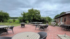 Una terrazza esterna con tavoli rotondi e sedie in vimini, pavimento in mattoni e vista su un prato e alberi sotto un cielo nuvoloso.