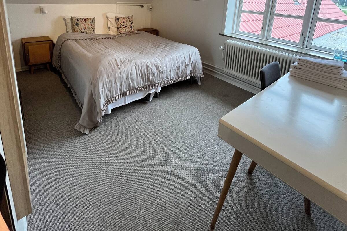 Une chambre à coucher avec un lit, un bureau, un radiateur et une fenêtre donnant sur un bâtiment au toit rouge.