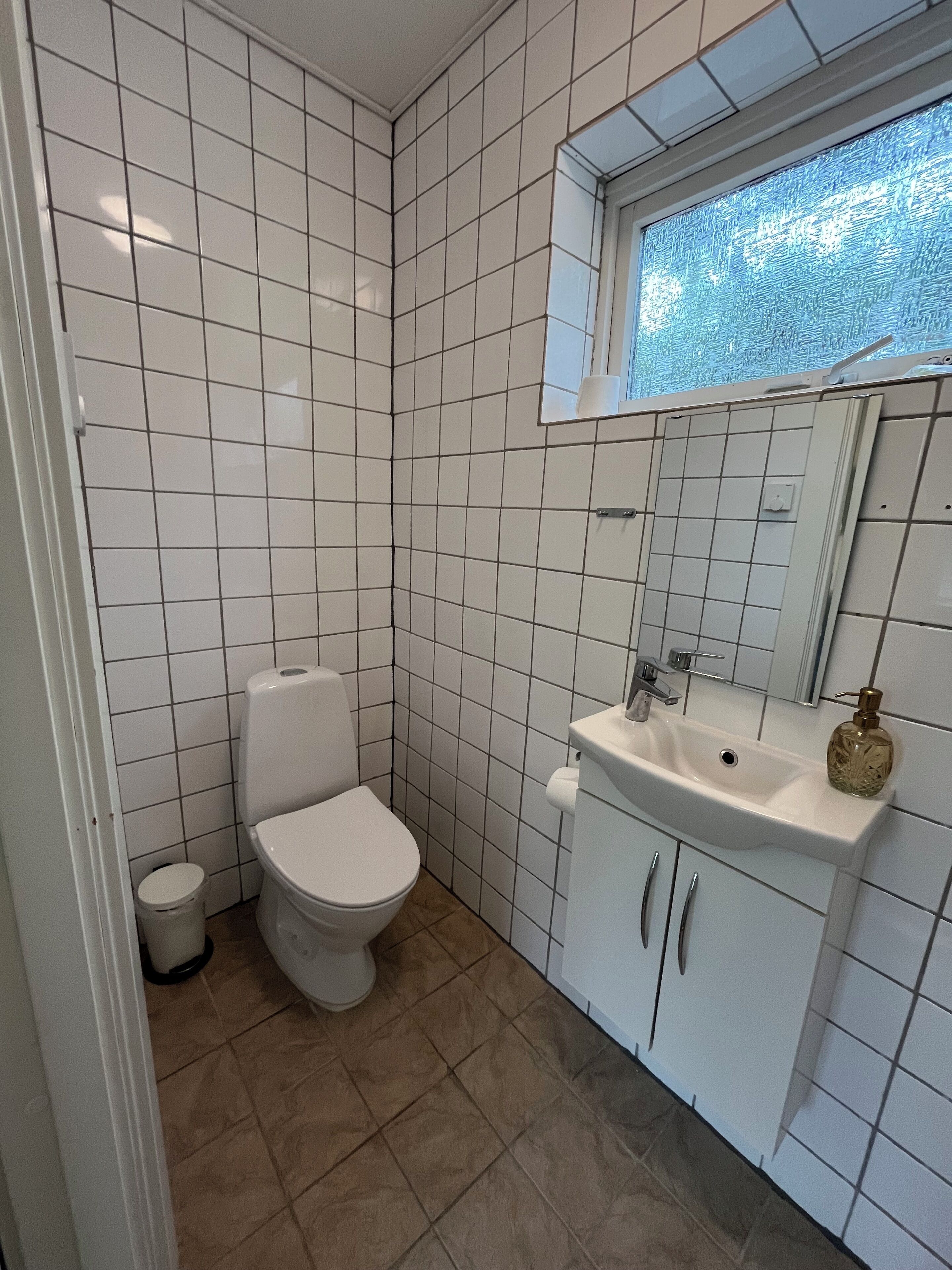 Een badkamer met witte betegelde wanden, een wit toilet, een wastafel met spiegel, en een raam boven de wastafel.