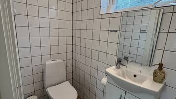 Een badkamer met witte betegelde wanden, een wit toilet, een wastafel met spiegel, en een raam boven de wastafel.
