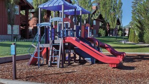 Un parque infantil con toboganes y estructuras para escalar, rodeado de árboles y un edificio al fondo.