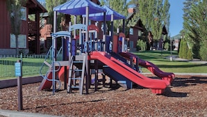 Un parque infantil con toboganes y estructuras para escalar, rodeado de árboles y un edificio al fondo.