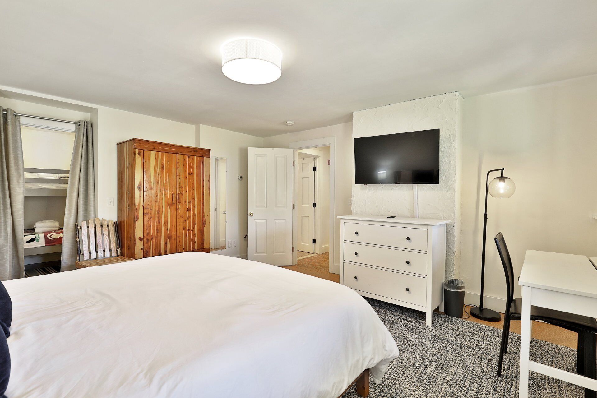 Un dormitorio con una cama grande, un armario de madera, un tocador blanco, un televisor montado en la pared y un escritorio con una lámpara.