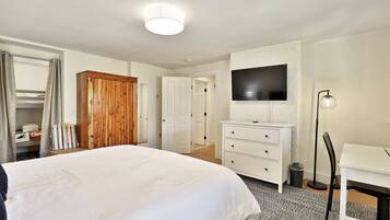 Un dormitorio con una cama grande, un armario de madera, un tocador blanco, un televisor montado en la pared y un escritorio con una lámpara.