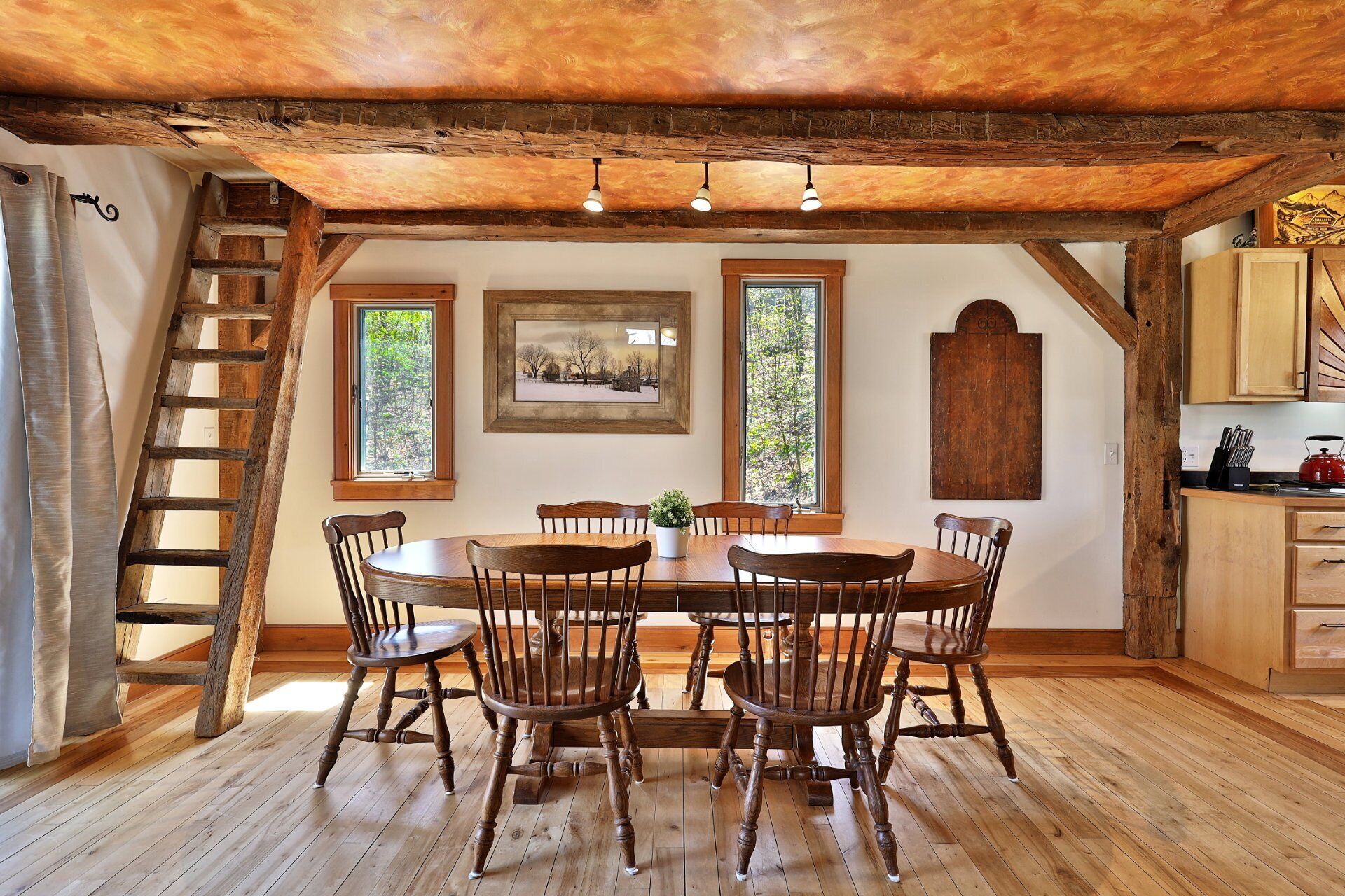 Une salle à manger rustique avec une table et des chaises en bois, une échelle et une vue sur l’extérieur par les fenêtres.