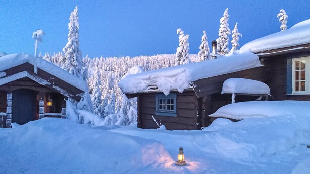 En stuga av trä med snötäckta tak och en upplyst lykta i förgrunden.