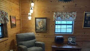 L’intérieur chaleureux d’un chalet avec un fauteuil gris, des murs en bois, une fenêtre ornée de rideaux à motifs floraux, un tableau encadré et une radio ancienne posée sur une table en bois.