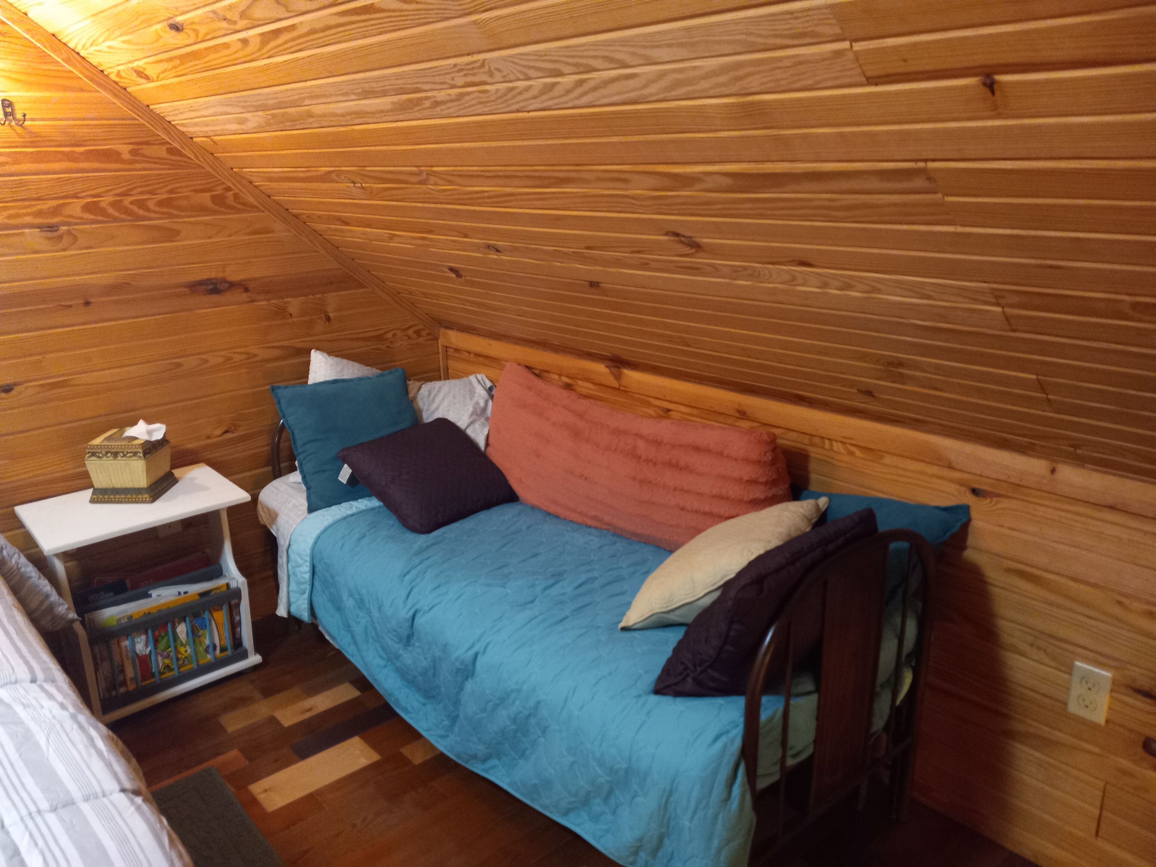 Ein kleines, gemütliches Zimmer mit holzgetäfelter, geneigter Decke, einem Einzelbett mit blauer Bettdecke und vielen Kissen, einem kleinen Nachttisch mit einer Schachtel und Taschentüchern sowie einem Babybett mit einer bunten Spielkiste.