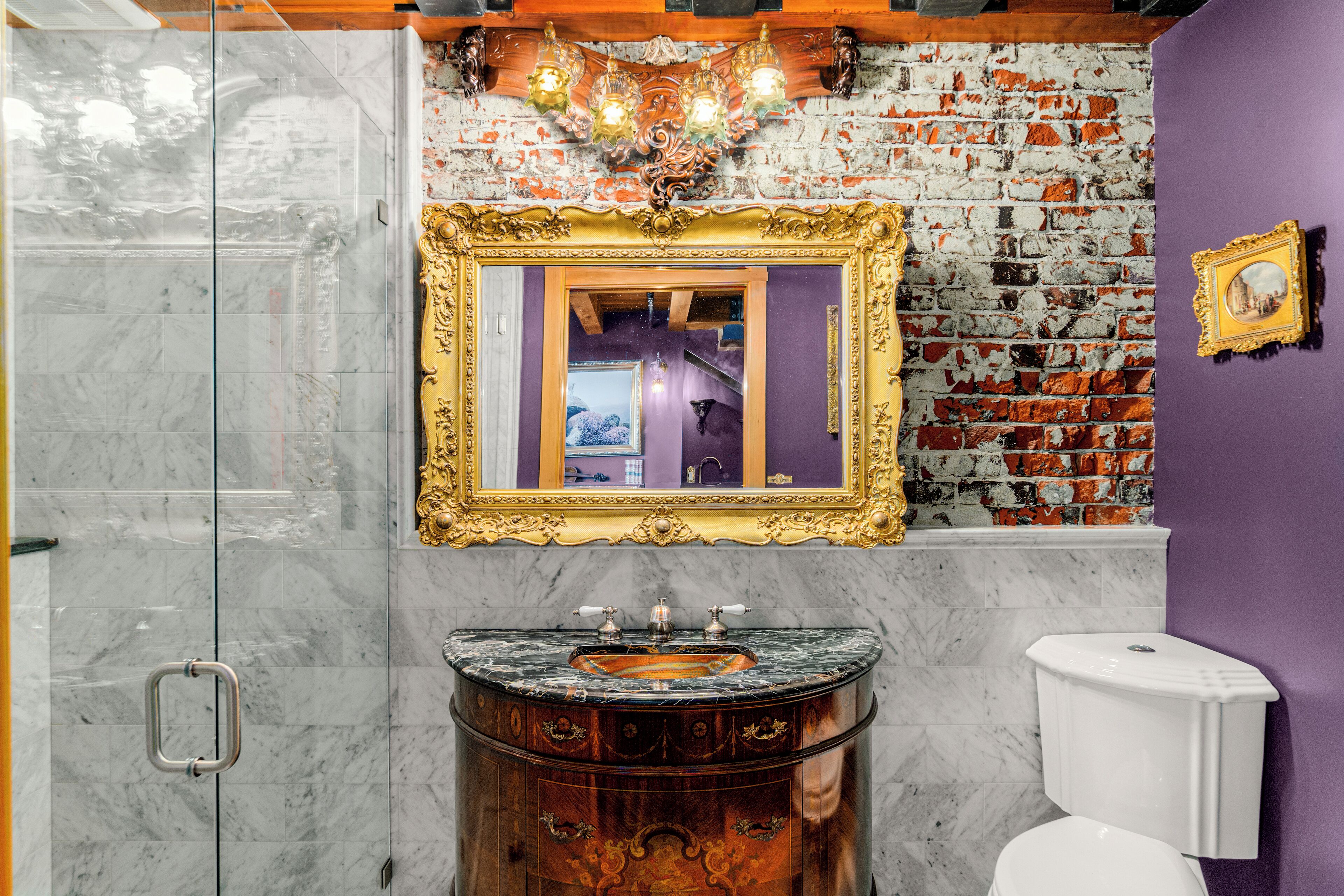 Een badkamer met een marmeren wastafel, een spiegel met gouden lijst, een wit toilet en een bakstenen muur.