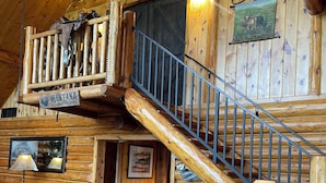 A rustic cabin interior with a wooden staircase, a leather sofa, a dining table with a horse sculpture, and a sign indicating Montana as the Treasure State.