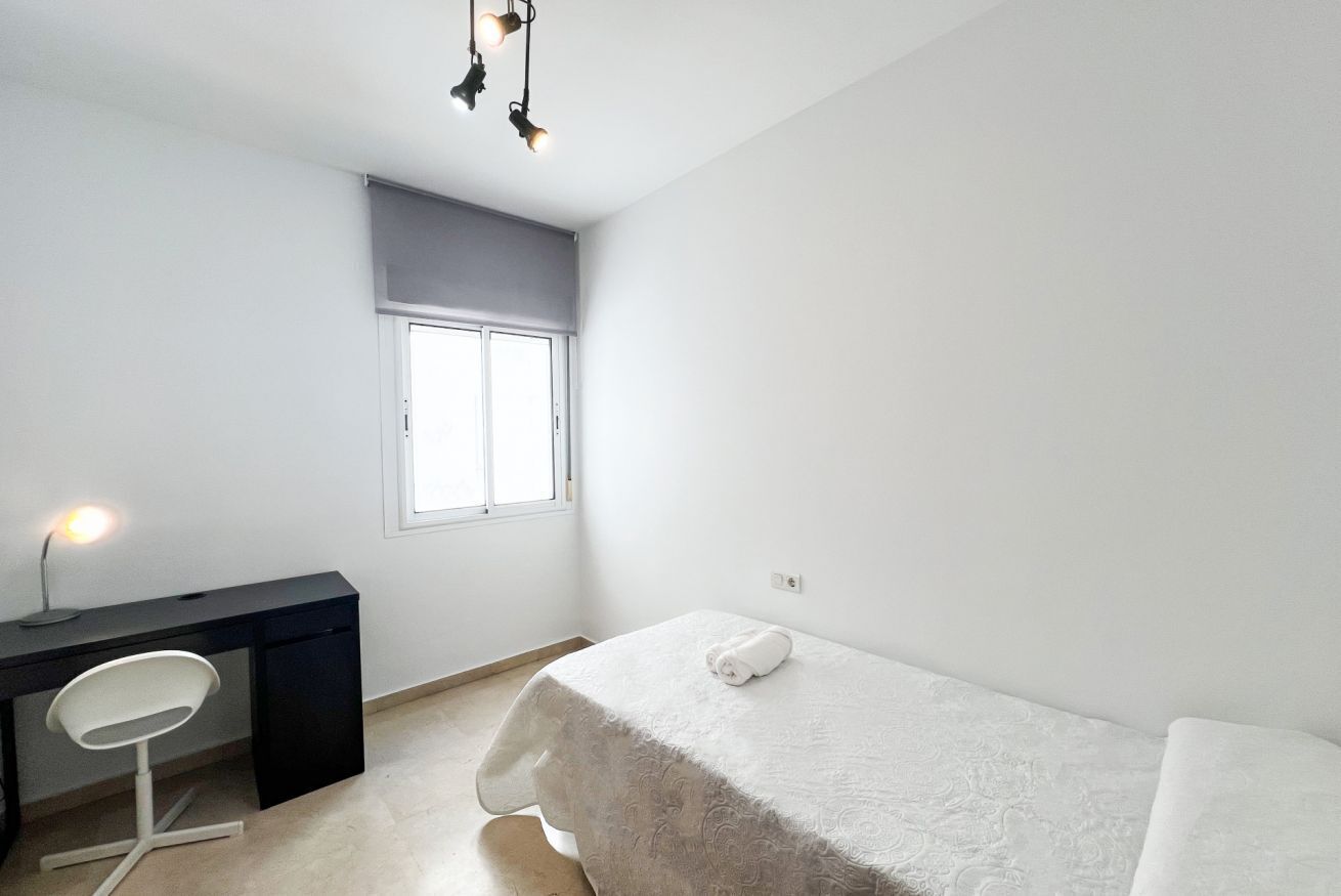 A small, clean room with a bed, a desk, and a chair. There is a window with a white blind, a lamp on the desk, and a towel on the bed.